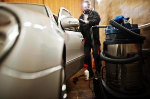 Man in uniform and respirator, worker of car wash center, cleaning car interior with hot steam cleaner. Car detailing concept. photo