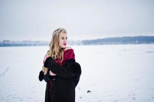 Elegance blonde girl in fur coat and red evening dress posed at winter snowy day. photo