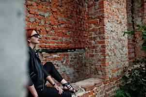 Red haired stylish girl in sunglasses wear in black, against abadoned place with brick walls. photo