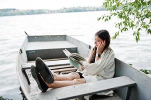 Portrait of an attractive woman wearing black polka dots dress, white shawl and glasses reading a book in a boat on a lake. photo