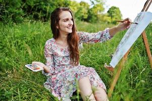 retrato de una hermosa joven feliz con un hermoso vestido sentada en el césped y pintando sobre papel con acuarelas. foto