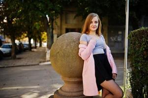 Young blonde girl in black skirt and pink coat posed at sunny day on street. photo