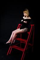 retrato de estudio de una chica rubia con maquillaje original en el cuello y tatuaje en el muslo, vestida con un vestido negro de fondo oscuro, sentada en una escalera roja. foto