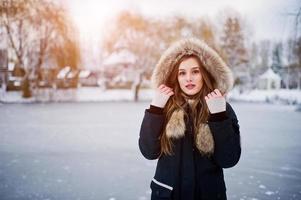 Beautiful brunette girl in winter warm clothing. Model on winter jacket against frozen lake at park. photo