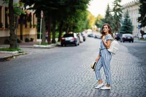 Portrait of a beautiful model in striped overall posing with hat and a backpack on a street with trees in a town. photo