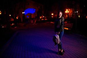 Night portrait of girl model wear on glasses, jeans and leather jacket, with backpack in hands, against blue lights garland of city street. photo