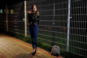 Night portrait of girl model wear on jeans and leather jacket against iron fence. photo