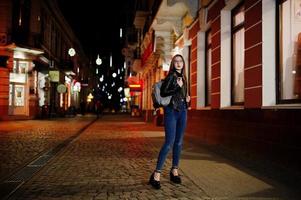 Night portrait of girl model wear on glasses, jeans and leather jacket, with backpack , against lights of city streets. photo