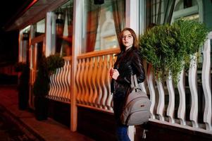 Night portrait of girl model wear on glasses, jeans and leather jacket, with backpack , against city streets. photo