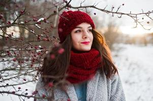 Portrait of gentle girl in gray coat , red hat and scarf near the branches of a snow-covered tree. photo