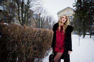 Elegance blonde girl in red evening dress and fur coat at streets of city in winter day. photo