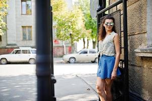 Curly stylish girl wear on blue jeans skirt, blouse and sunglasses. Portrait on streets of city. photo