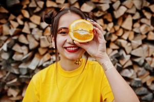 jovencita divertida con maquillaje brillante, vestida con camisa amarilla, sostenga una pieza de naranja sobre fondo de madera. foto