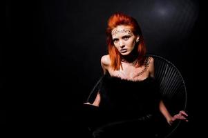 Fashion model red haired girl with originally make up like leopard predator isolated on black. Studio portrait on chair. photo