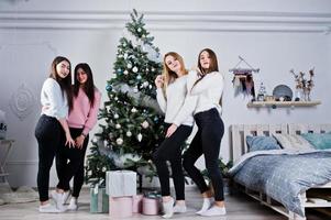 Four cute friends girls wear on warm sweaters, black pants against new year tree with christmas decoration at white room. photo