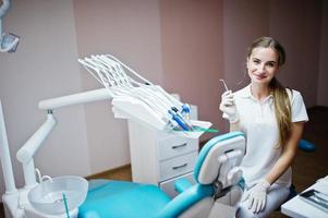 Good-looking female dentist posing with some dental instruments in her hand in white coat in a modern well-equipped cabinet. photo