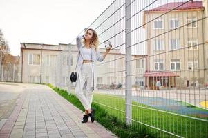 Stylish curly blonde model girl wear on white posing against fence. photo