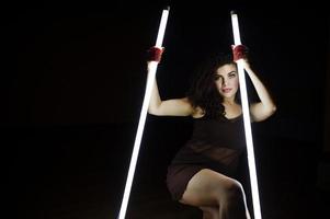 Curly brunette plus size model with two long led lamp at black room. photo