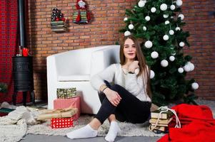 ropa de niña linda en suéteres cálidos, pantalones negros contra el árbol de año nuevo con decoración navideña en el estudio. foto