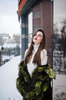 Brunette girl in green fur coat at street of city against house with large windows at winter. photo