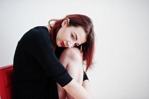 Red haired girl on black dress tunic sitting on red chair against white wall at empty room. photo