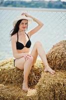 Portrait of a gorgeous girl in black bikini swimsuit posing on the hay bale with a hat by the lake. photo