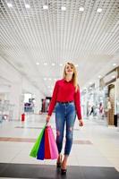 retrato de una mujer joven impresionante en blusa roja, jeans casuales rasgados y tacones altos posando con bolsas de compras en el centro comercial. foto