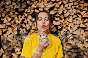 joven chica divertida con maquillaje brillante, use una camisa amarilla soplando burbujas de jabón contra un fondo de madera. foto