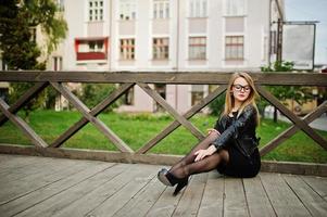 elegante chica rubia vestida con chaqueta de cuero negro posando en las calles de la ciudad sentada en madera. foto