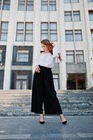 Portrait of a fabulous young successful woman in white blouse and broad black pants posing on the stairs with a huge white building on the background. photo