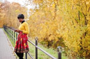 niña afroamericana con vestido amarillo y rojo en el parque de otoño dorado con tableta en las manos. foto