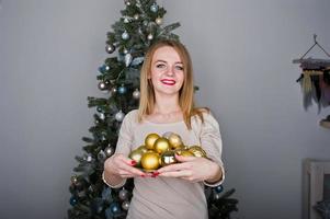 linda chica rubia con vestido beige contra el árbol de año nuevo con bolas doradas de año nuevo en el estudio. felices vacaciones. foto