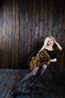 Elegante blonde girl wear on fur coat sitting on chair at studio against wooden background. photo