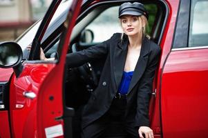 Portrait of beautiful blonde sexy fashion woman model in cap and in all black, blue corset, with bright makeup near red city car. photo