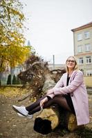 chica rubia con gafas y abrigo rosa, túnica negra sentada en un árbol cortado. foto