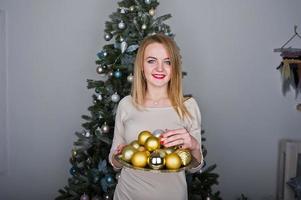 Cute blonde girl on beige dress against new year tree with with golden new year balls at studio. Happy holidays. photo