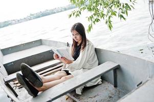 Portrait of an attractive woman wearing black polka dots dress, white shawl and glasses reading a book in a boat on a lake. photo