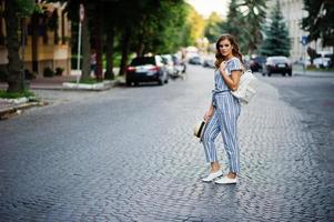Portrait of a beautiful model in striped overall posing with hat and a backpack on a street with trees in a town. photo