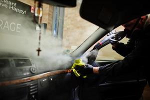 Man in uniform and respirator, worker of car wash center, cleaning car interior with hot steam cleaner. Car detailing concept. photo