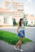 Curly stylish girl wear on blue jeans skirt, blouse and listening music from headphones phone. Portrait on streets of city. photo