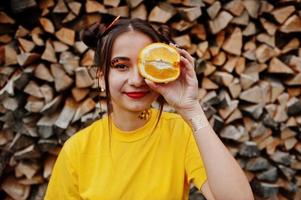 Young funny girl with bright make-up, wear on yellow shirt hold picece of orange against wooden background. photo
