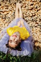 Young funny girl with bright make-up, like fairytale princess, wear on yellow shirt and violet skirt lying against wooden background. photo
