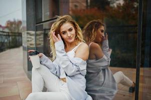 Stylish curly blonde model girl wear on white with cup of coffee at hand posing against large window. photo