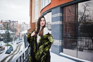 chica morena con abrigo de piel verde en la calle de la ciudad contra la casa con grandes ventanales en invierno. foto