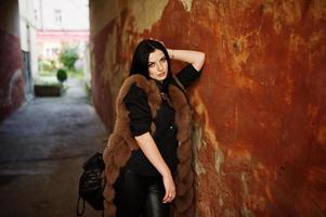 Fashion outdoor photo of gorgeous sensual woman with dark hair in elegant clothes and luxurious fur coat at old street with grunge walls.