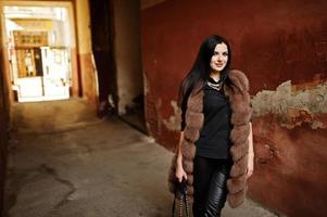 Fashion outdoor photo of gorgeous sensual woman with dark hair in elegant clothes and luxurious fur coat at old street with grunge walls.