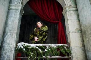 Brunette girl in green fur coat against old arch with columns and red curtains. photo