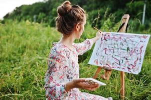 Portrait of a gorgeous happy young woman in beautiful dress sitting on the grass and painting on paper with watercolors. photo