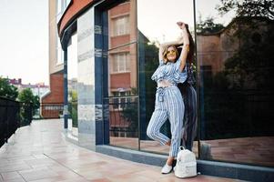 Portrait of a gorgeous young woman in striped overall and yellow sunglasses posing with white leather backpack on a balcony of a modern glass building in town. photo