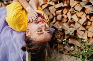 Young funny girl with bright make-up, like fairytale princess, wear on yellow shirt and violet skirt lying against wooden background. photo